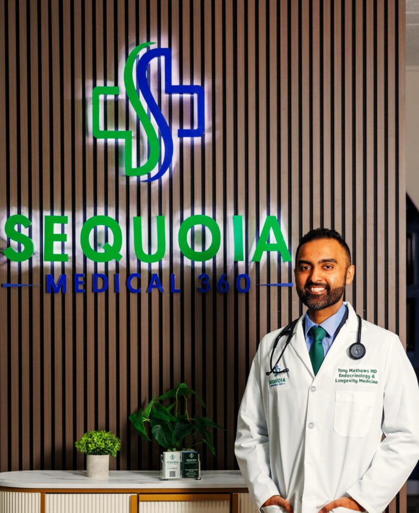 Dr. Tony Mathews, MD, standing in front of the Sequoia Medical 360 logo wall wearing a white coat and stethoscope inside the clinic’s modern Bronxville office.